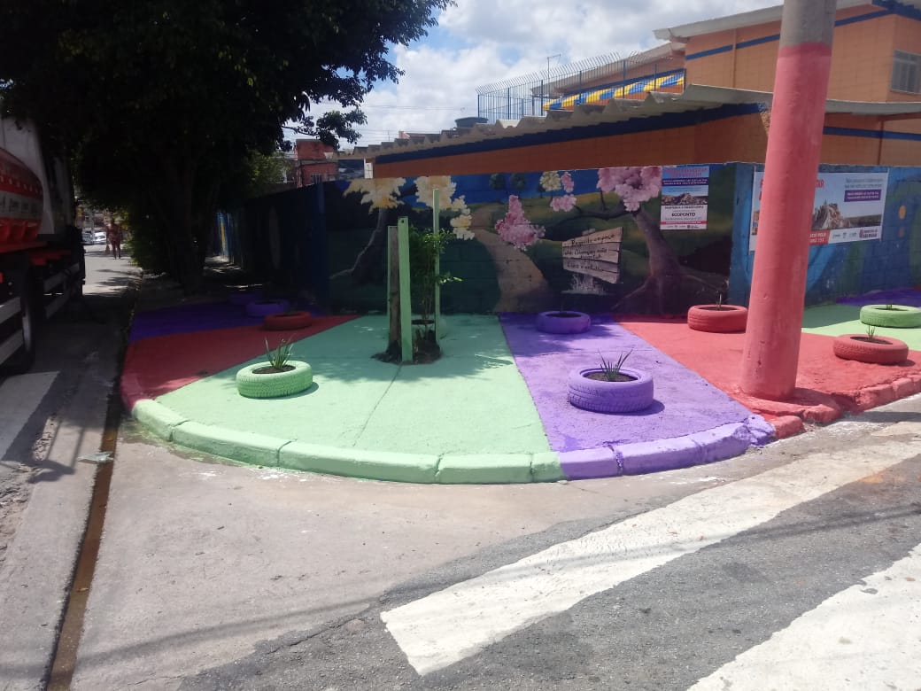 esquina com calçada larga pintada com faixas verde, vermelha e lilás. à beira da rua, pneus também pintados servem de floreiras. 